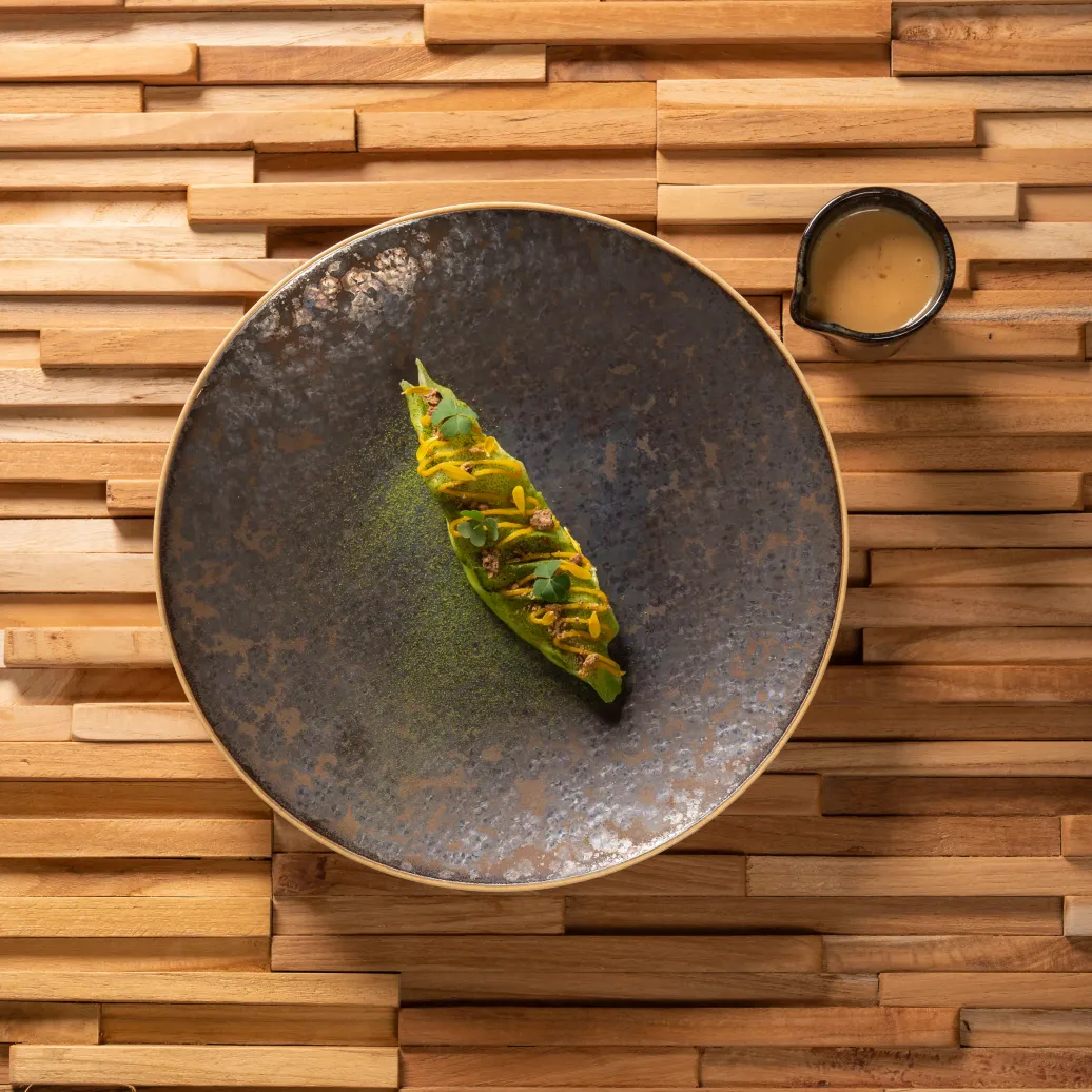Un plat contenant un légume vert entouré de poudre verte, posé sur un mur en bois avec un petit bol à côté