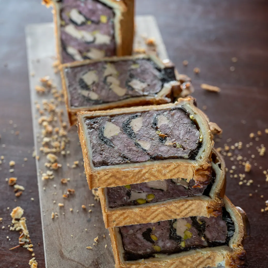 Un gâteau rectangulaire coupé en tranches, avec une croûte dorée et une garniture pourpre, posé sur une planche en bois avec des miettes autour