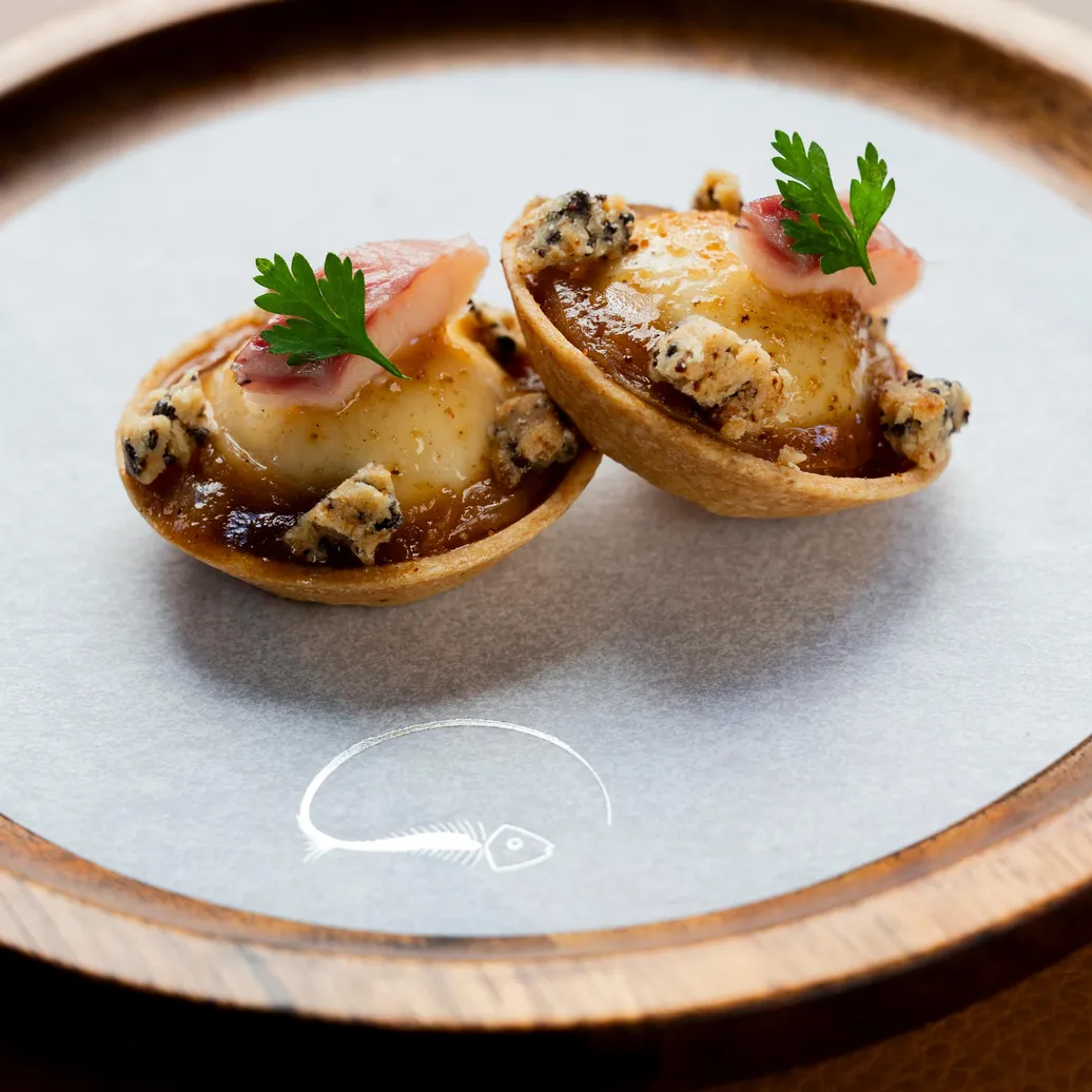 Deux petits pâtés garnis de crème et de morceaux de viande, décorés de fines tranches de radis et de feuilles de persil, présentés sur une assiette grise à bord en bois avec un motif de poisson