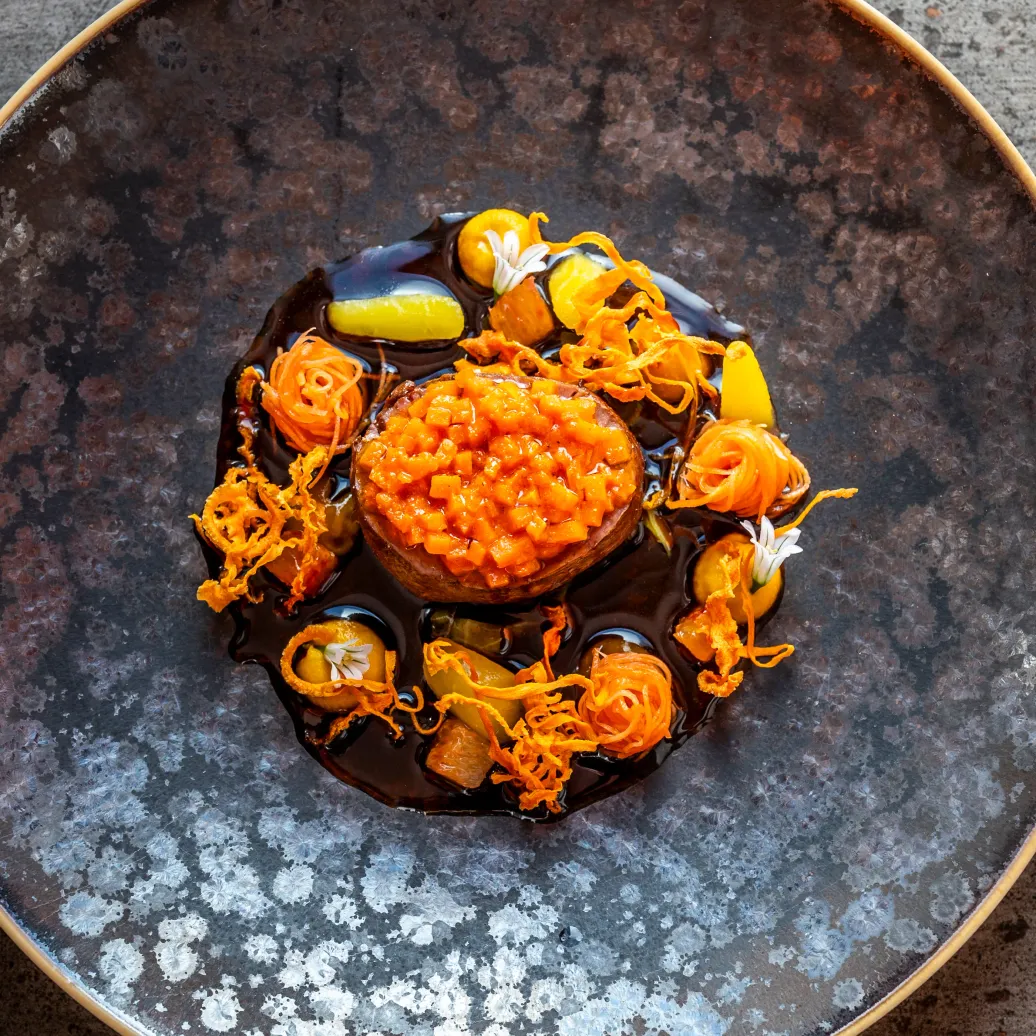 Un plat gourmet avec une préparation orange au centre, entouré de garnitures variées et de sauce, servi dans un bol métallique