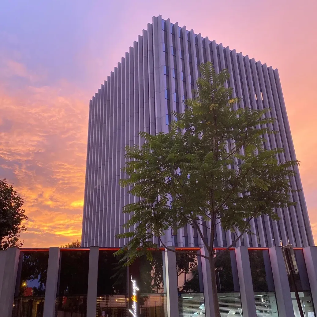 Bâtiment moderne à façade verticale striée avec des arbres devant, sous un ciel de coucher de soleil aux teintes orangées et roses