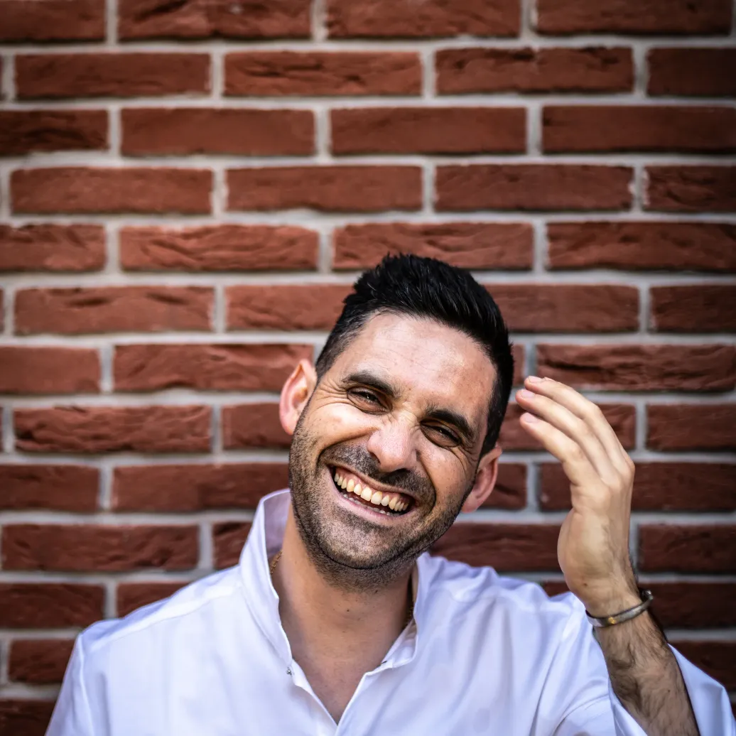 Homme souriant devant un mur de briques, portant une chemise blanche avec un nom brodé