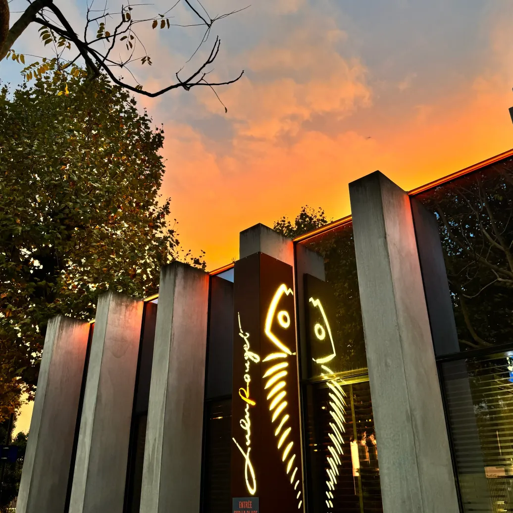 Bâtiment moderne avec une enseigne lumineuse en néon jaune et un panneau indiquant "Entrée sur la place" au coucher du soleil