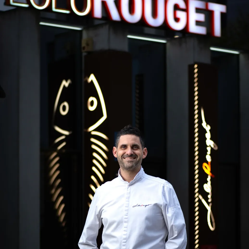 Un homme souriant en chemise blanche et jean devant l'enseigne lumineuse Lulu Rouge