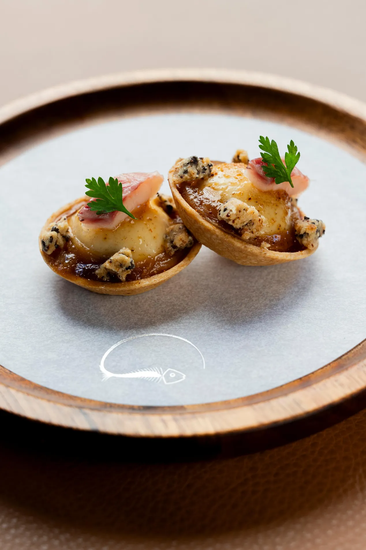 Deux petits pâtés garnis de crème et de morceaux de viande, décorés de fines tranches de radis et de feuilles de persil, présentés sur une assiette grise à bord en bois avec un motif de poisson