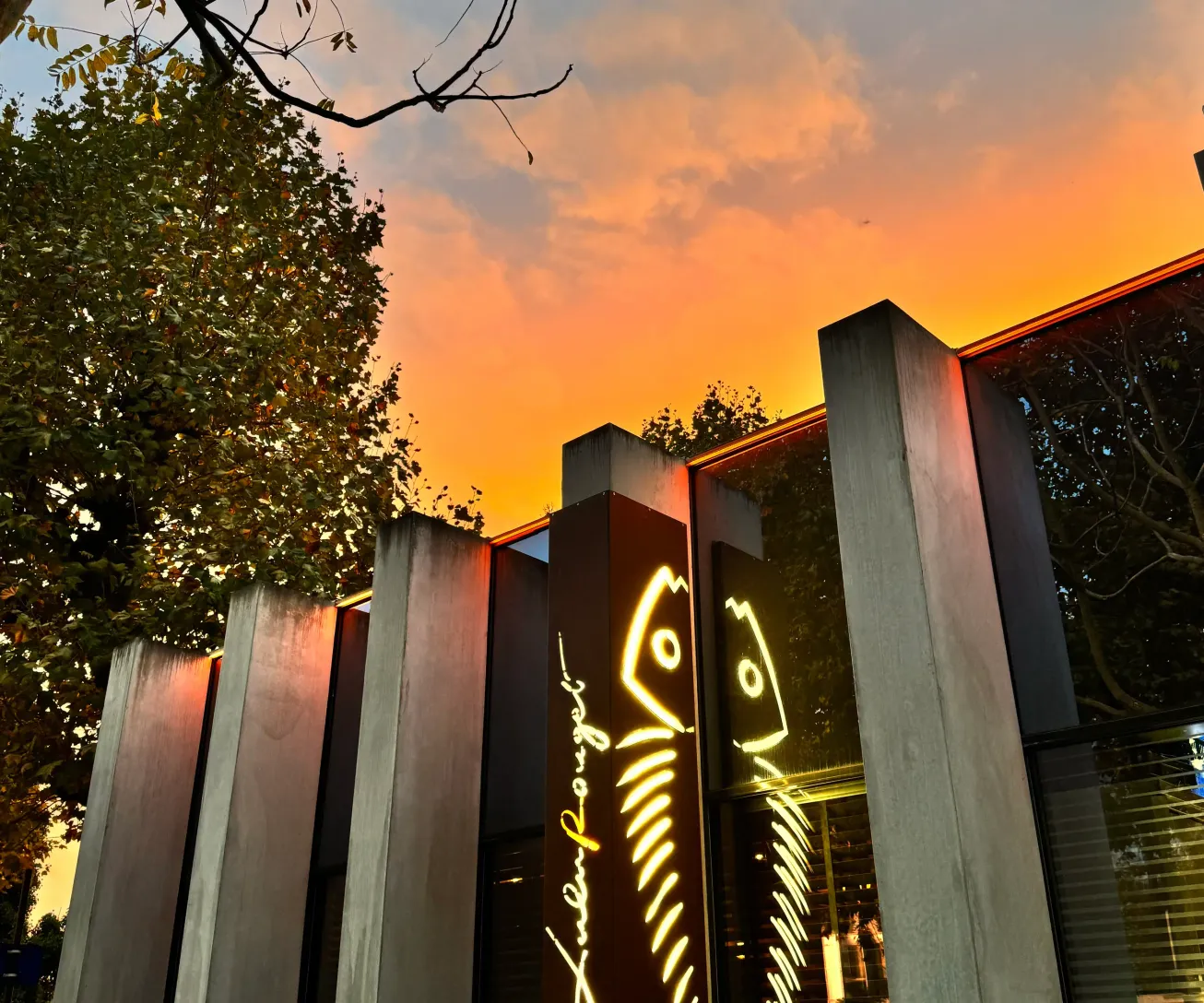 Bâtiment moderne avec une enseigne lumineuse en néon jaune et un panneau indiquant "Entrée sur la place" au coucher du soleil