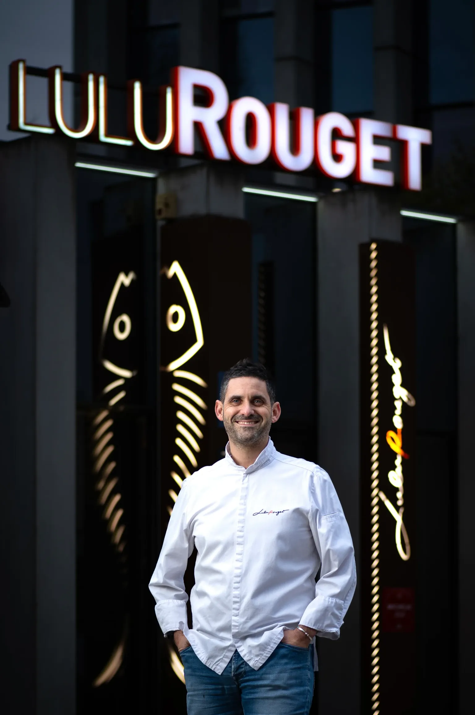 Un homme souriant en chemise blanche et jean devant l'enseigne lumineuse Lulu Rouge