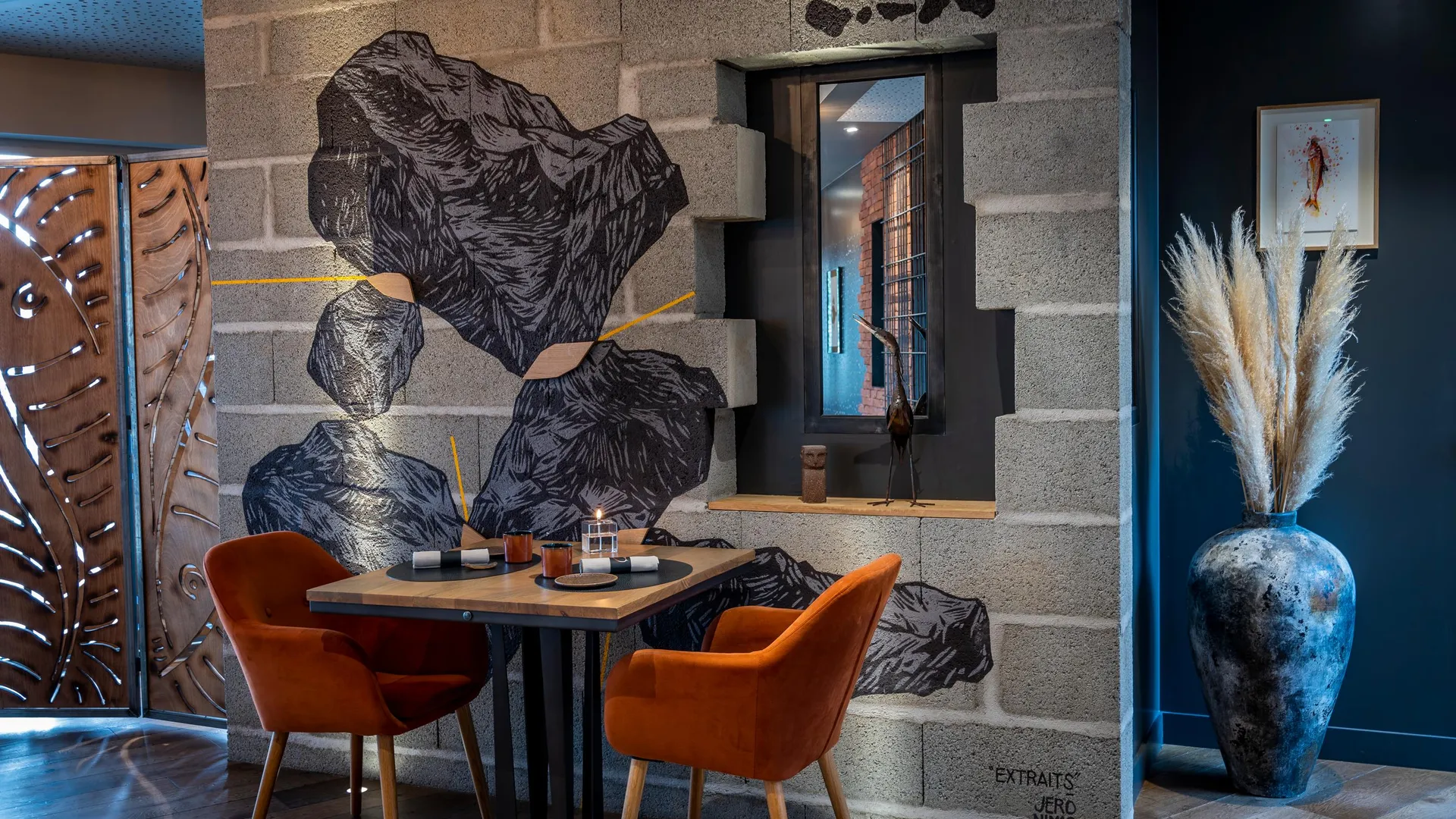 Table avec deux chaises orange dans un intérieur décoré de grandes feuilles murales et d'un vase avec des herbes sèches