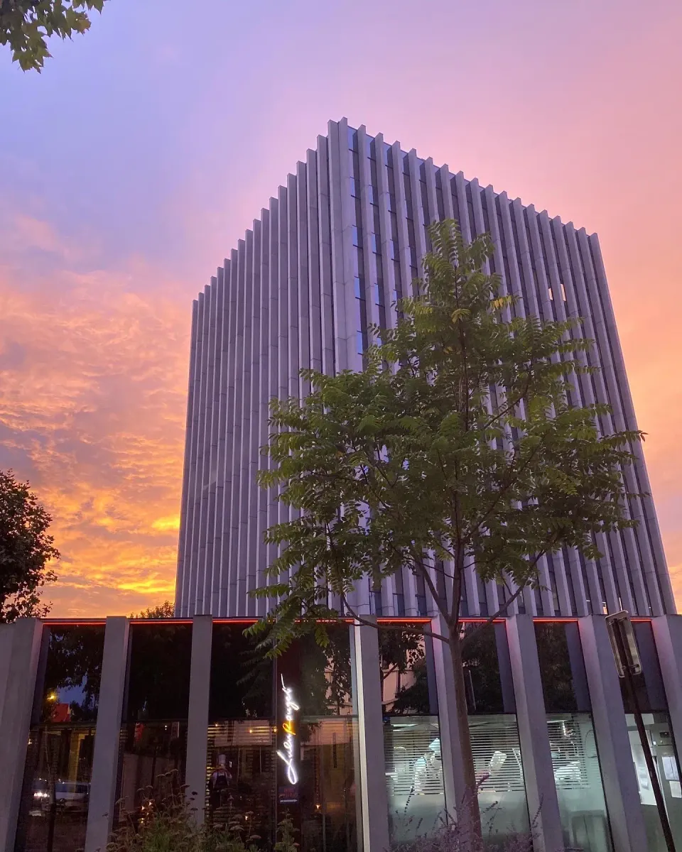 Bâtiment moderne à façade verticale striée avec des arbres devant, sous un ciel de coucher de soleil aux teintes orangées et roses