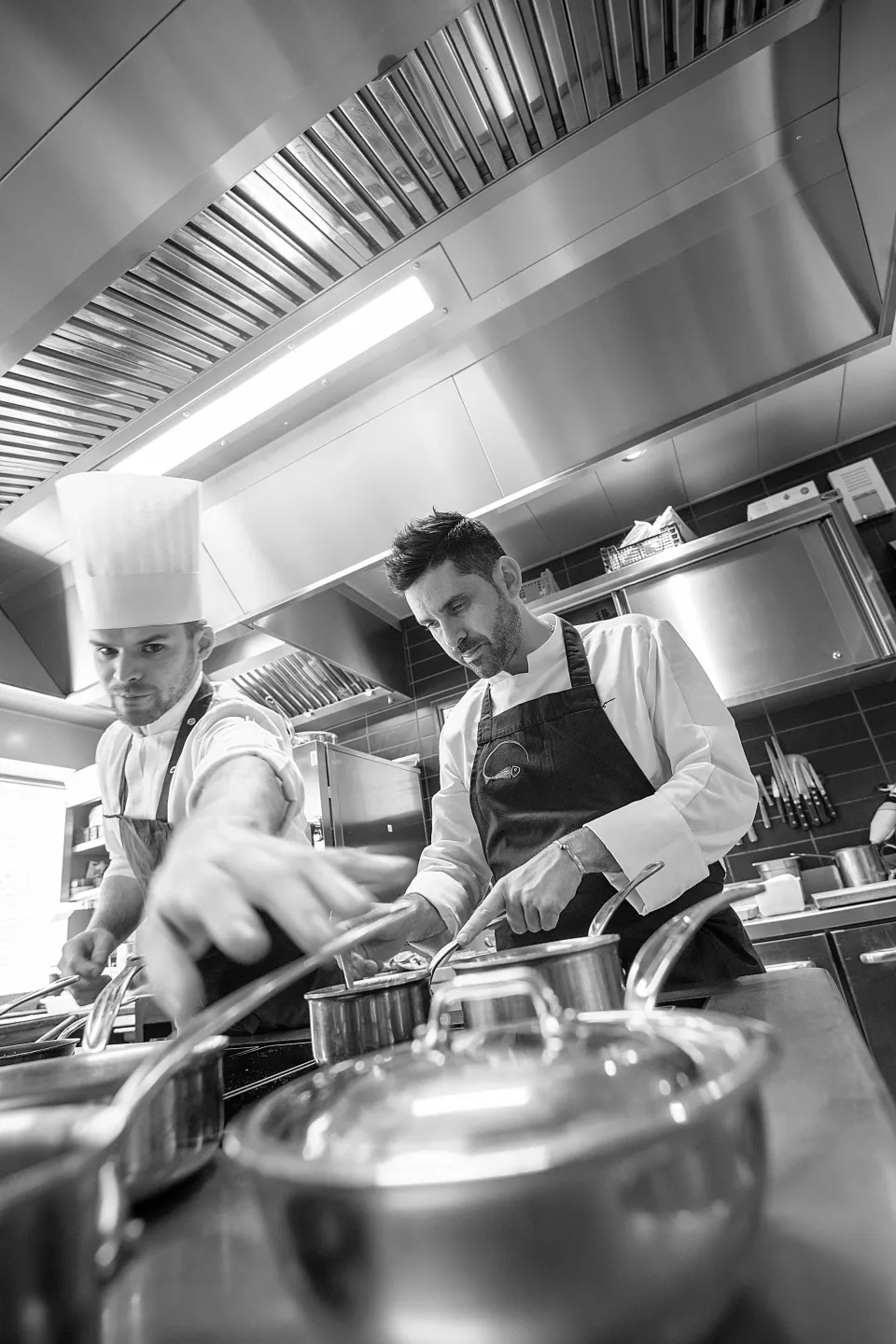 Deux chefs cuisiniers en uniforme travaillent dans une cuisine professionnelle, préparant des plats à l'aide de divers ustensiles et casseroles